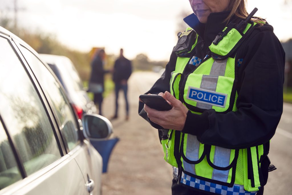 A police officer checks a mobile phone beside a car during an investigation.